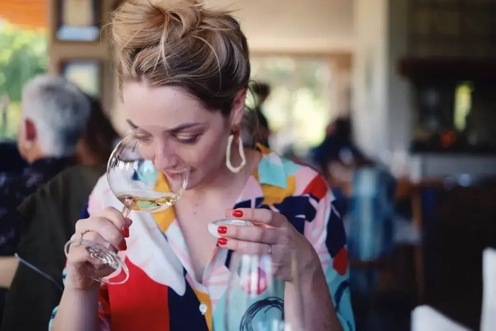 Een vrouw met een wijnglas, gekleed in een kleurrijk, patroonrijk shirt, zit aan een tafel in een bruisend restaurant.